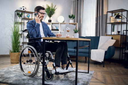 Disabled young man talking on smartphone and working on wireless laptop at home. Male freelancer sitting in wheelchair and using modern gadgets.の写真素材