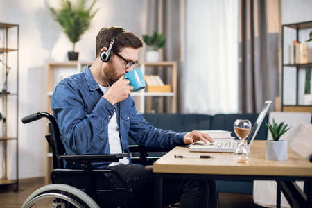 Side view of young man with special need using wireless laptop for remote work at home. Bearded guy sitting in wheelchair, drinking coffee and typing on computer.の写真素材