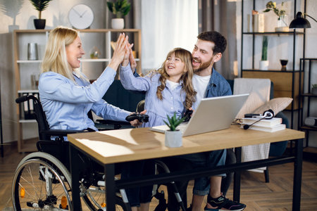 Young woman in wheelchair giving high five to cute daughter and handsome husband. Happy family...