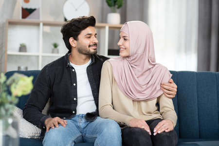 Home, family and relationship concept. Close up portrait of happy smiling arabian couple, handsome bearded man and charming woman in hijab, hugging while sitting on sofa at home and looking each otherの写真素材