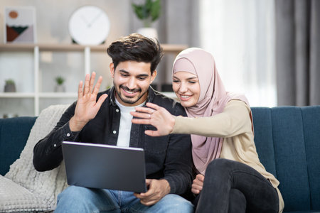 Laughing cheerful young muslim couple sitting on a couch with a laptop at home, waving while having video call talking with relatives or friends. Family, technology, wifi internet concept.の写真素材