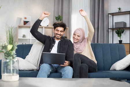 Cheerful young Muslim couple, holding hands up in victory gesture, celebrating success. Arabian...