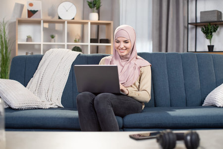 Online education and distance work from home. Happy Arabic Muslim girl in headscarf, sitting on blue...