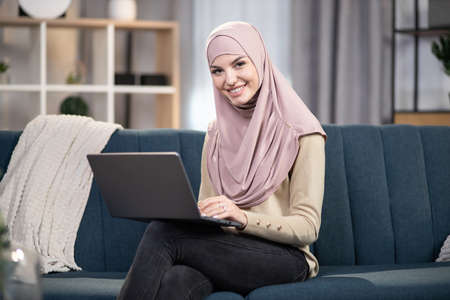 Gorgeous young Muslim woman wearing hijab, using laptop at home, shopping online, working or studying. Muslim woman using laptop sitting on sofa at home living room.の写真素材