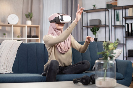 Pleasant young Muslim girl wearing VR technology glasses and touching air by hands, using 3D viewer...