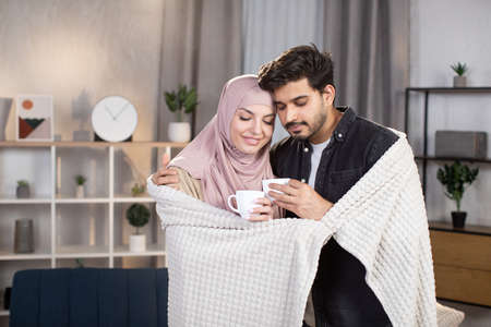 Family, cozy moments and love concept. Cute Arabian Muslim couple relaxing under blanket at home in the living room and drinking hot coffeeの写真素材