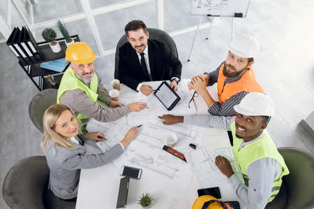 Designers, architects and engineers gathering at modern office for creating project of new building. Partners sitting at table and working with blueprints. People of various ages, gender and races.の写真素材