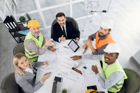 Top view of five diverse people in suits and helmets sitting at office and showing thumbs up....