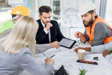 Multiracial office workers in suits and helmets having conference about construction project. Men and women cooperating to create new modern high-rise building.の写真素材