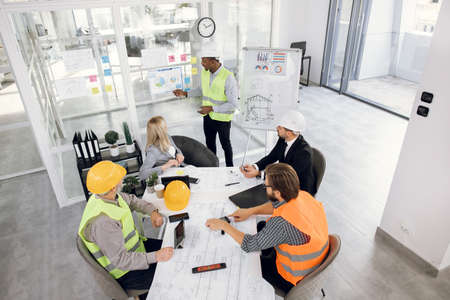 Top view of multi ethnic people in formal wear and helmets having working conference at office. Five diverse workers analysing graphs and chart of new building plan.の写真素材