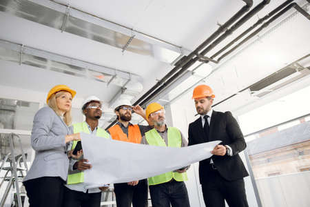 Four bearded man and one beautiful woman analysing blueprints of new building while standing indoors. Multiracial colleagues wearing protective helmets on construction site.の写真素材