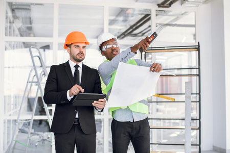 Group of two multi ethnic men in protective helmets inspecting industrial plan. International...