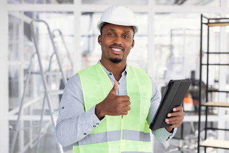 Happy african man in white construction hardhat and uniform showing thumb up and smiling on camera....