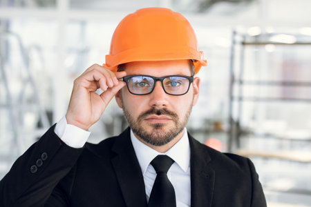 Close up portrait of handsome confident experienced bearded man, engineer or architect in orange helmet, wearing eyeglasses and looking at camera, standing at counstruction site indoorsの写真素材