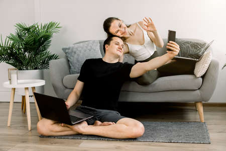 Happy sporty couple using smartphone and laptops for workout at home, having fun, sitting in lotus position on mat and sofa, relaxing after morning exercises, having video call with friends.の写真素材