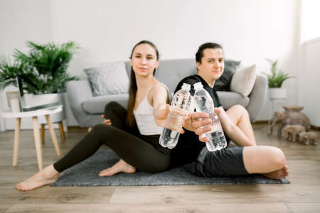 Sporty young man and woman, sitting on the floor back to back, resting after home workout, showing water in plastic bottles to camera. Water balance concept. Focus on the bottles.の写真素材