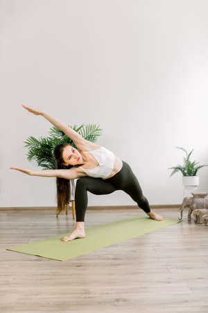Calmness and relax, woman happiness and yoga balance concept. Young beautiful woman practicing yoga indoors at modern light yoga hall or at home, standing on green mat in Floating Triangle pose.の写真素材