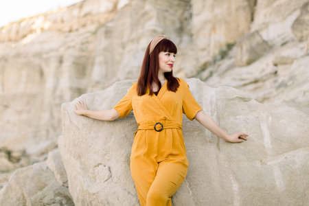 Summer fashion portrait of young beautiful red haired woman in yellow overalls and hair band, posing with arms outstretched, leaning on the stone in sand quarry outdoorsの写真素材