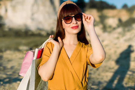 Close up portrait of elegant glamour young woman in sunglasses and yellow clothes, smiling with shopping bags outside, standing in front of beautifful sand quarry on summer sunset.の写真素材