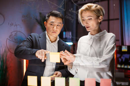 Caucasian and Asian colleagues standing in office behind the glass board. Blond lady puts stickers on board, focused man looking at the charts and planning project tasks. Teamwork creative approachの写真素材