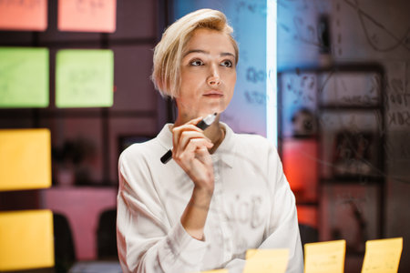 Pensive pretty businesswoman working on new project strategy, standing near glass board with sticky...