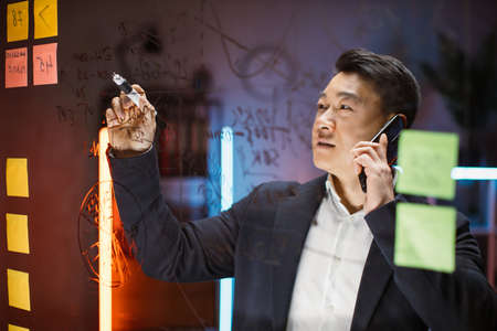 Focused Asian male ceo manager in formal wear writing on glass board with colorful sticky notes and talking phone, working with project in modern night office. Creative thinking, startup plan conceptの写真素材