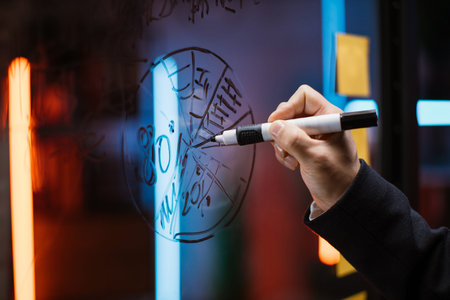 Close up side view of male hand drawing a pie marketing chart on glass office board with a black...