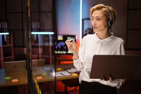 Mature professional female manager, wearing headset and using laptop, standing near glass wall in...