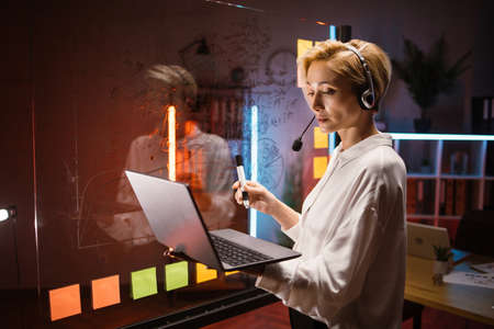 Concentrated blond businesslady writing tasks and ideas on glass wall with colorful stickers at office. Focused woman in headset using laptop for checking working progress, working with kanban boardの写真素材