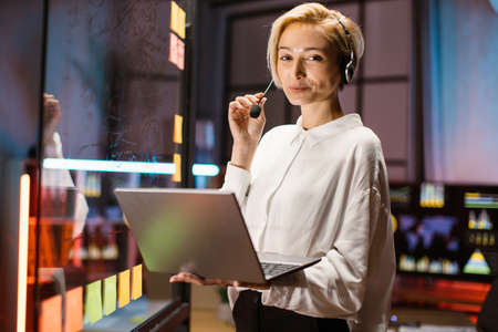 Portrait of pretty cheerful blond Caucasian executive businesswoman wearing headset using laptop, posing on camera with smile while standing near scrum kanban glass board at dark officeの写真素材