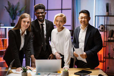Portrait of creative multiethnic business team standing together at the table with gadgets and papers and looking at camera. Multiracial business people together at the evening meeting.の写真素材