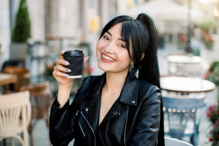 Beautiful young Asian girl with dark ponytail hair, dressed in black leather jacket, sitting at the table, drinking coffee and laughing. Summer, coffee break, people, urban lifestyle.の写真素材