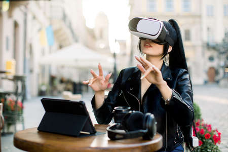 Pretty young smart Caucasian Asian businesswoman, sitting in outdoor cafe in front of tablet, working and watching virtual presentation in vr headset. City buildings on the background.の写真素材