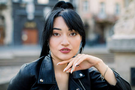 Close up of lovely sexy young mixed raced Asian woman in black leather jacket, posing on the street of old European city, on sunny day. Fashion portrait of pretty Asian girl.の写真素材