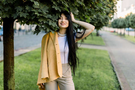 Portrait of pretty lovely Asian girl with long dark hair, posing on street on background of city park alley with green trees. Portrait of beautiful woman in trendy yellow blazer walking outdoors.の写真素材