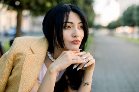 Young fashion brunette Asian business woman, dressed in elegant yellow jacket, sitting on the bench outdoor in the city, leaning her chin on hands and looking away. Close up urban portrait.の写真素材