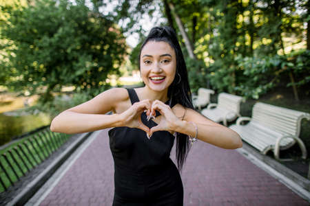 In love with world. Closeup portrait of modern smiling Chinese Asian girl showing heart shaped hands to camera, while standing outside in the summer green trees city park background.の写真素材