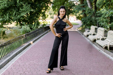 Full length portrait of young Asian woman model with ponytail hairstyle, smiling in urban park. Mixed raced girl wearing black trendy overalls and fashionable heels, posing on the park alley outdoors.の写真素材