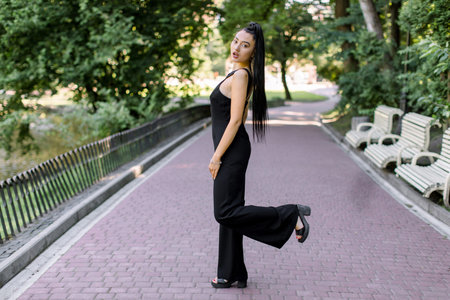 Outdoor portrait of beautiful sexy mixed raced Asian woman in black overalls, posing at park alley during sunset walk, with one leg raised up. Pretty brunette girl in elegant clothes at park.の写真素材