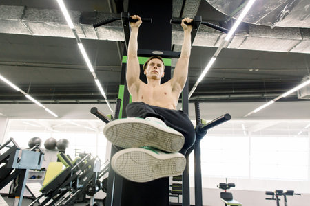Handsome shirtless man doing pull ups in the L-sit position on horizontal bar at the gym....