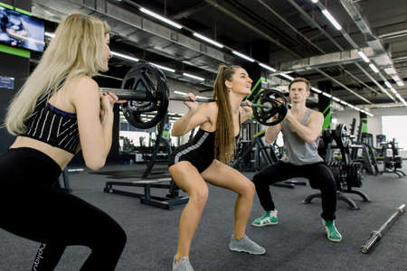 Fit young woman lifting barbell looking focused, working out in a gym with trainers.の写真素材