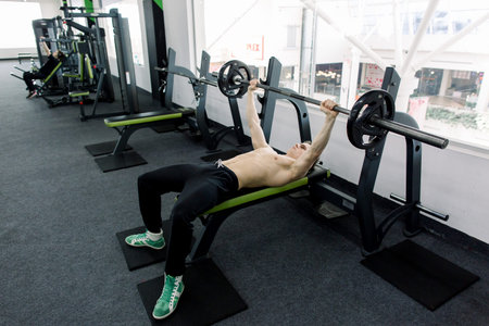 Young muscular Caucasian man lifting barbell on the bench in the gym
