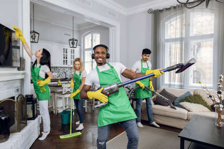 African man listening music in headphones and dancing like guitarist with modern vacuum cleaner. Three multiracial cleaners in uniform wiping, washing and vacuuming on background.の写真素材