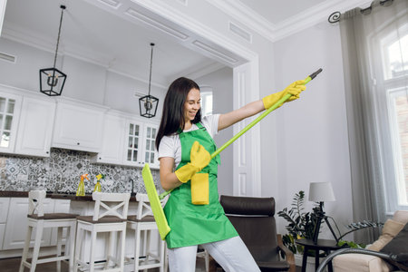 Smiling young woman with brown hair in green apron and yellow gloves playing on mop like on guitar....