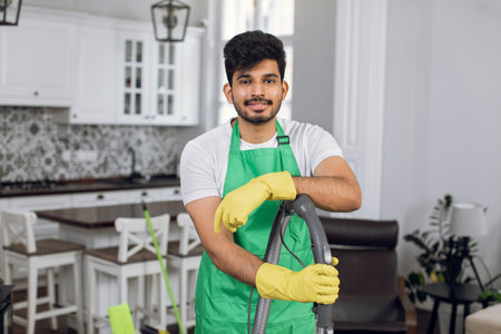 Handsome indian man in uniform and rubber gloves standing indoors at bright room. Happy male cleaner with modern vacuum smiling on camera.の写真素材
