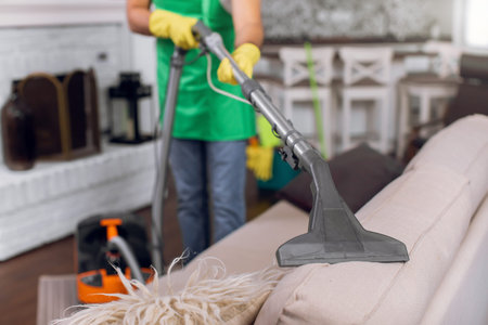 Close up of male cleaner in uniform and gloves vacuuming cozy sofa at bright room. Cleaning process...