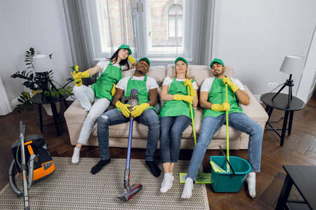 Tired multiracial group of cleaners in uniform, hat and gloves relaxing together on couch after general cleanup of bright house. Modern cleaning service.の写真素材