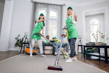 Two female janitors in uniform using modern vacuum for cleaning room. Their colleagues, multiracial men sitting on couch and using tablet on background. Addiction from gadgets.の写真素材