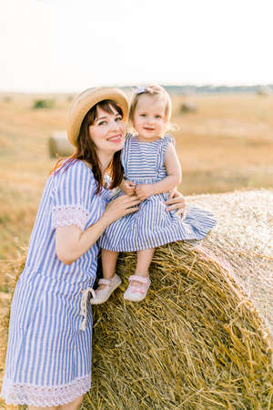 Close up portrait of cute little baby girl sitting on hay stack bale and posing to camera together with her charming young mother. Mommy and daughter having fun walking in wheat field on sunny day.の写真素材