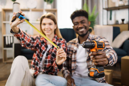 Blur background of multi ethnic couple in casual clothes showing measure tape and electric drill on camera. Cheerful family using tools for repairing and assembling furniture at new house.の写真素材
